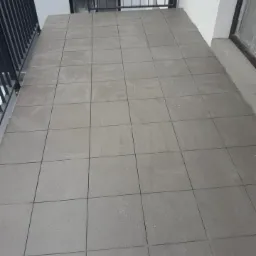 Szare, kwadratowe płytki ceramiczne ułożone na balkonie z metalową balustradą w nowoczesnym budynku.