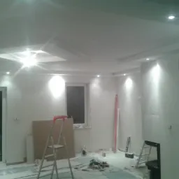 Wnętrze w trakcie remontu: ściany po szpachlowaniu, sufit z oświetleniem punktowym i dekoracyjnymi wgłębieniami, drabina oparta o ścianę, narzędzia na podłodze zabezpieczonej folią.