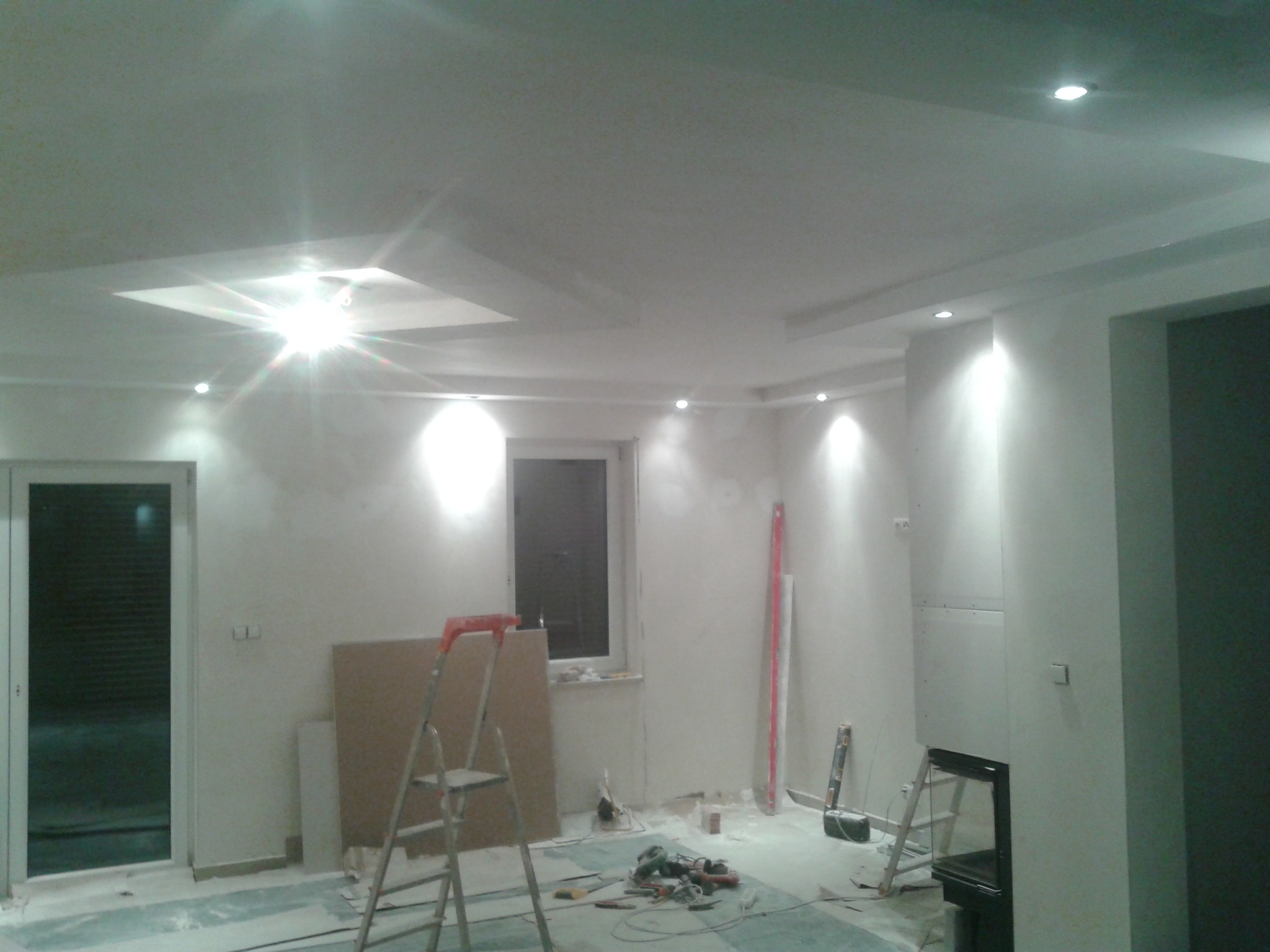 Wnętrze w trakcie remontu: ściany po szpachlowaniu, sufit z oświetleniem punktowym i dekoracyjnymi wgłębieniami, drabina oparta o ścianę, narzędzia na podłodze zabezpieczonej folią.
