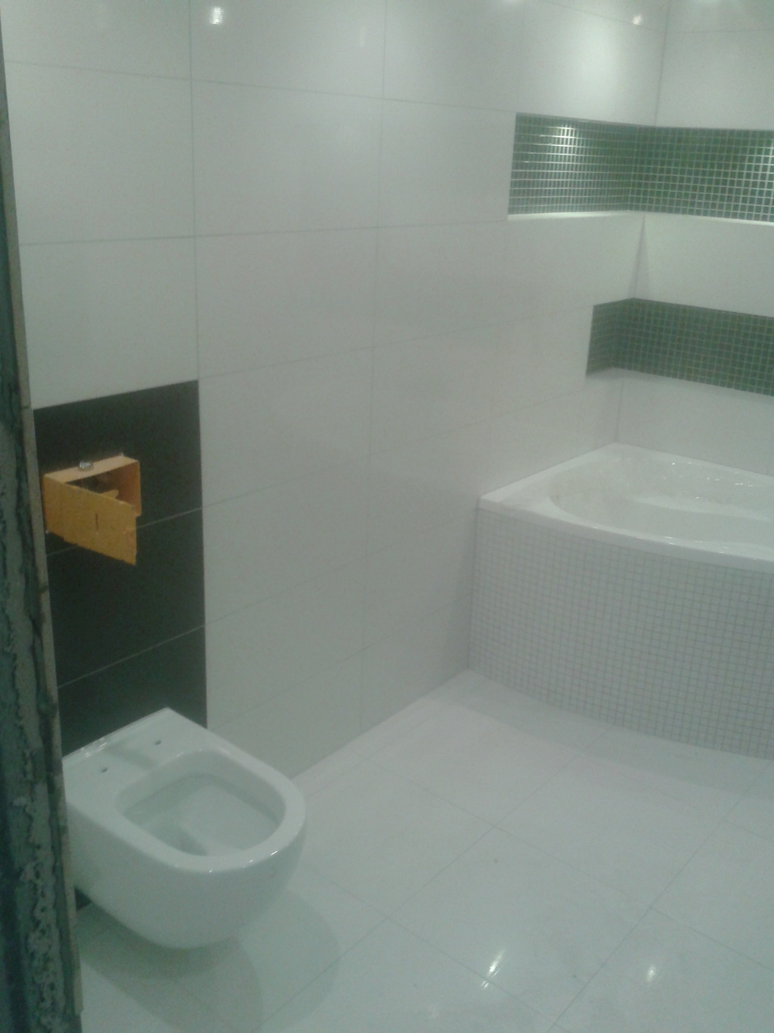 Biała łazienka z częściowo wykończonymi ścianami, widoczna instalacja podtynkowego WC, obudowana mozaiką wanna oraz wnęki z zieloną mozaiką na ścianach.