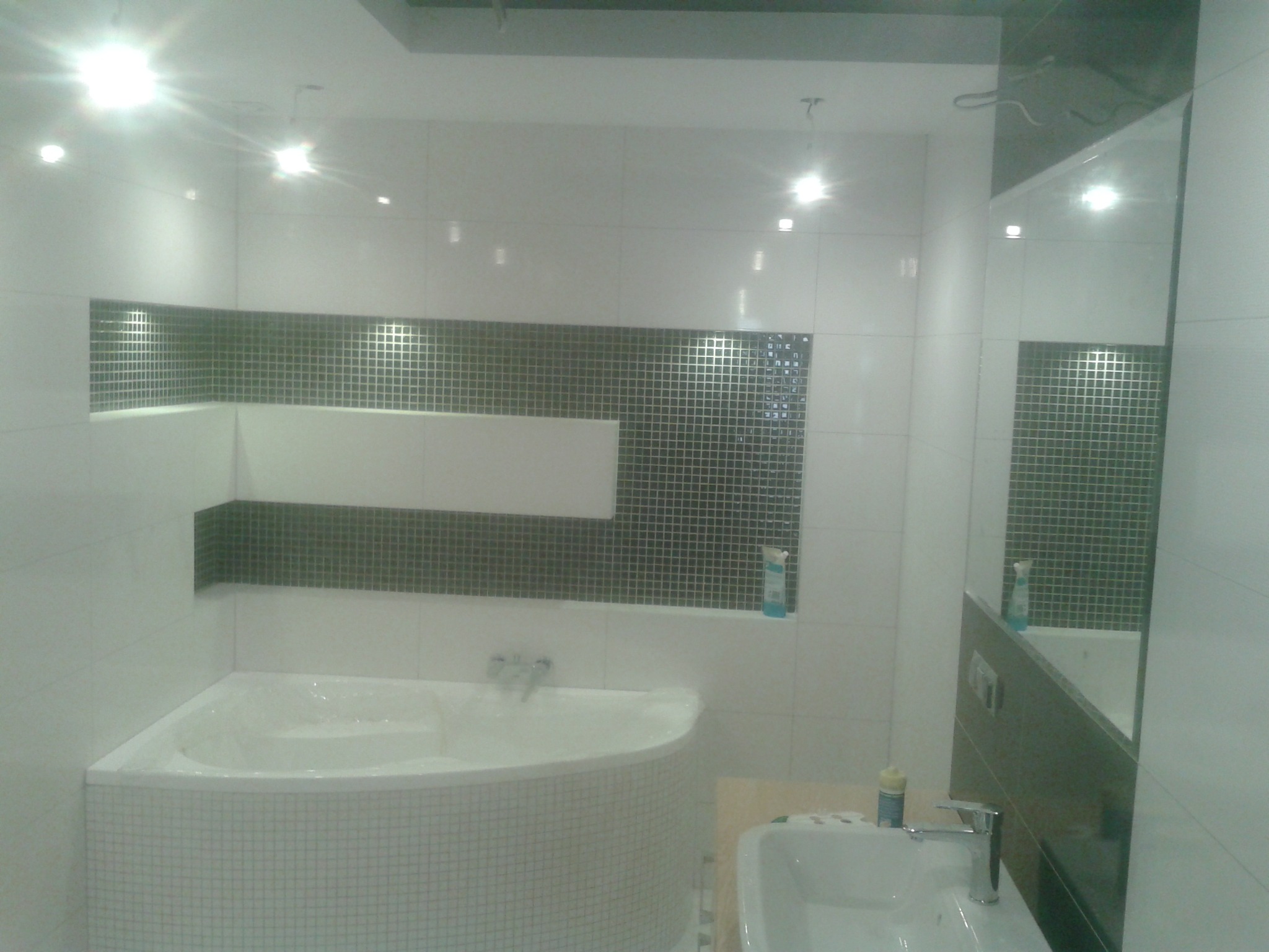 Nowoczesna łazienka z narożną wanną obudowaną mozaiką, umywalką z chromowaną baterią i lustrem, akcentowana szarą mozaiką wnękową i punktowym oświetleniem sufitowym.