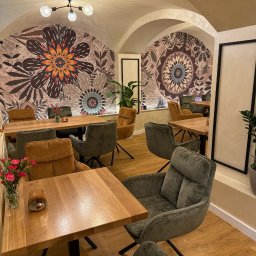 DESIGNKING KINGA KUPIJAJ - Wnętrze kawiarni z drewnianymi stołami, tapicerowanymi krzesłami i dekoracyjnymi muralami kwiatowymi na ścianach. Oświetlenie sufitowe i rośliny doniczkowe dopełniają aranżację.