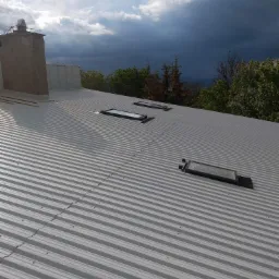 Szary, blaszany dach z trzema oknami dachowymi i kominem na tle burzowego nieba.