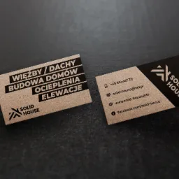 Dwie wizytówki firmy budowlanej Solid House na ciemnoszarym tle. Jedna wizytówka w kolorze kraftowym z czarnym tekstem: więźby/dachy, budowa domów, ocieplenia elewacji i logo. Druga wizytówka...