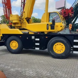 Żółty dźwig samochodowy z wysuniętymi podporami na placu, widoczne inne dźwigi w tle.