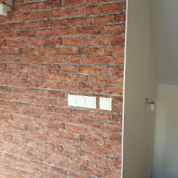 Ściana pokryta tapetą imitującą czerwoną cegłę z widocznymi trzema białymi włącznikami światła.