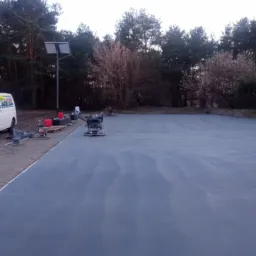 Świeżo wylana, szara posadzka zacierana mechanicznie z widocznymi dwoma zacieraczkami mechanicznymi, w tle las i dwa busy firmy.