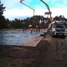 Betonowanie podłoża na boisku sportowym z użyciem pompy do betonu z logo firmy buszrem.pl, widoczni pracownicy w odblaskowych ubraniach, podłoże zabezpieczone folią.