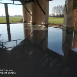 Świeżo wylana, gładka posadzka w surowym budynku gospodarczym z widokiem na pole, odblaski na powierzchni, widoczne drewniane elementy konstrukcyjne.