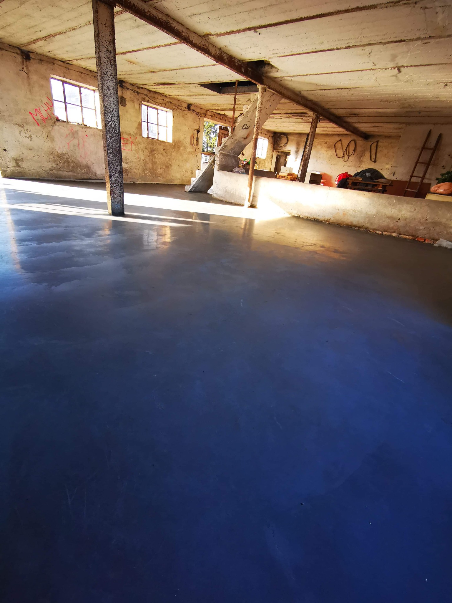 Świeżo wylana, gładka, ciemnoszara podłoga w surowym, industrialnym wnętrzu z betonowym stropem, drewnianymi belkami, oknami i widocznymi śladami po remoncie na ścianach.