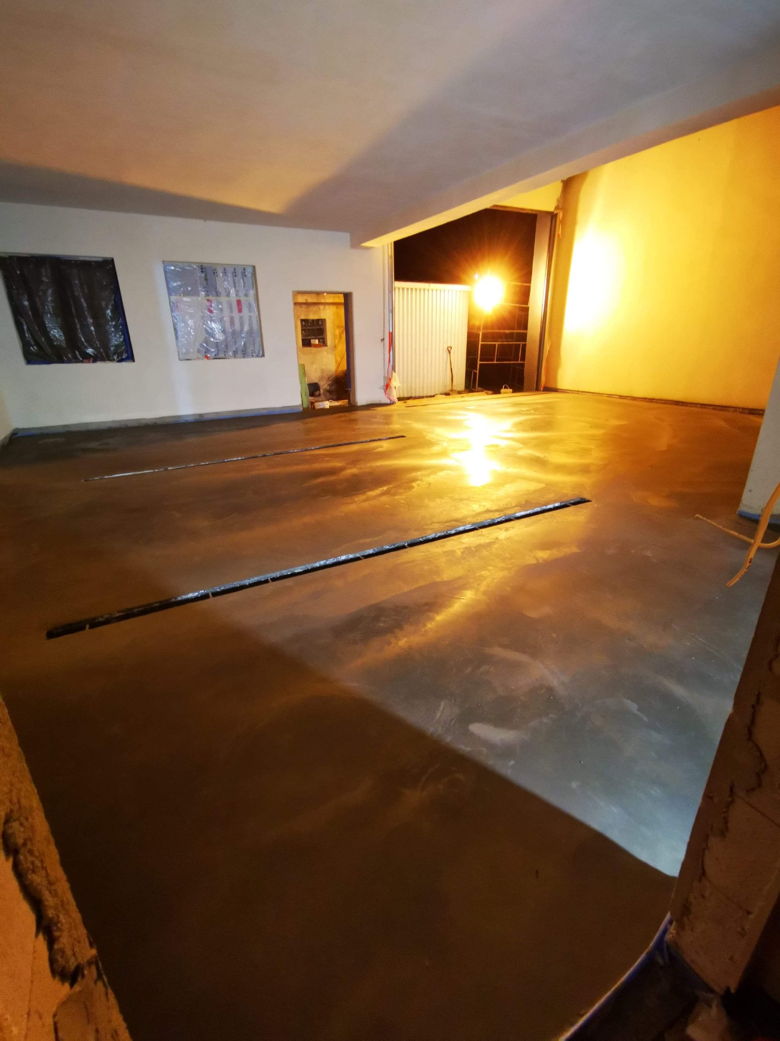 Świeżo wylana, błyszcząca posadzka przemysłowa z widocznymi szczelinami dylatacyjnymi, zabezpieczone okna folią malarską, oświetlenie halogenowe.
