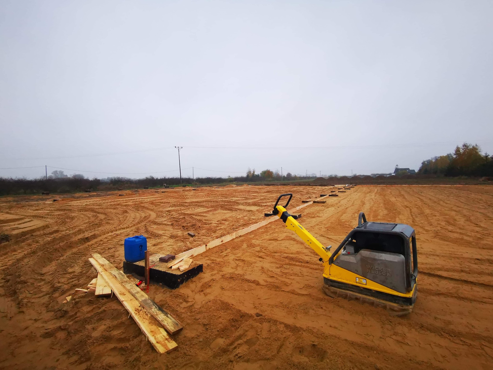 Żółta zagęszczarka Wacker Neuson ubija piasek na placu budowy pod fundamenty, widoczne drewniane szalunki i niebieski kanister, pochmurne niebo w tle.