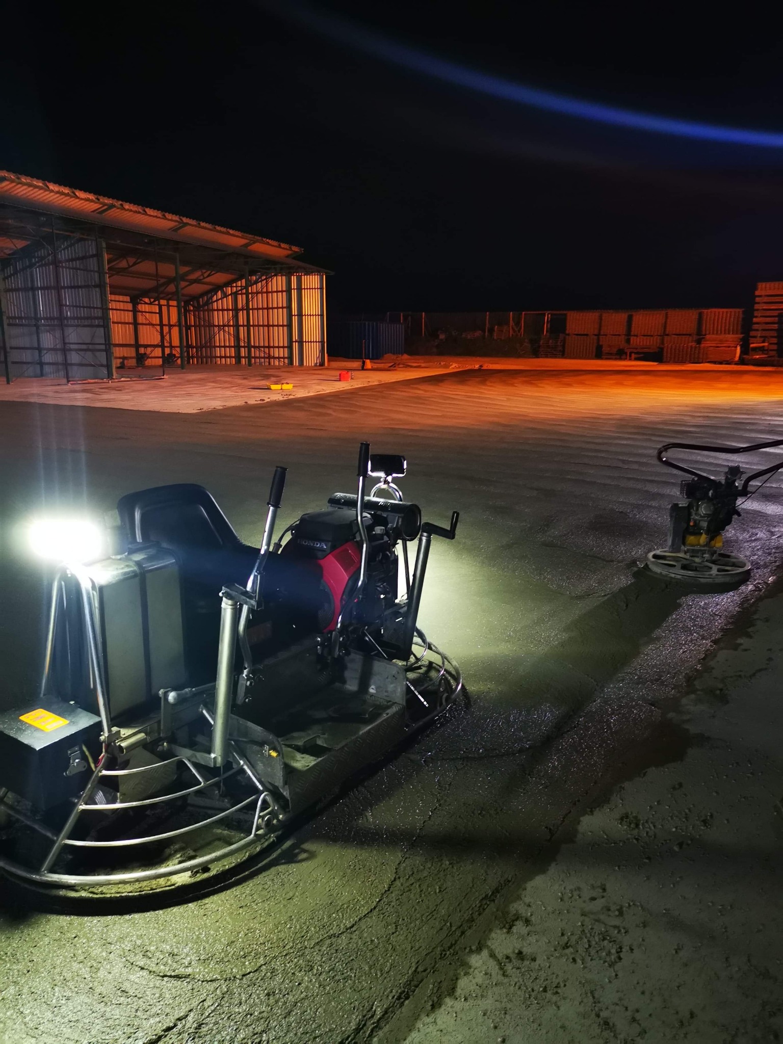 Dwie zacieraczki mechaniczne do betonu pracują nocą na dużej powierzchni świeżo wylanego betonu w hali przemysłowej oświetlonej pomarańczowym światłem.