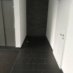 Korytarz z ciemnymi, prostokątnymi płytkami na podłodze i ścianą wyłożoną czarnym kamieniem dekoracyjnym, oświetlony z góry.