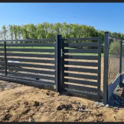 Nowoczesna, grafitowa brama wjazdowa z poziomymi profilami, otwierana furtka obok, w tle zielone drzewa i ogrodzenie panelowe na betonowej podmurówce, teren wokół w trakcie prac ziemnych.