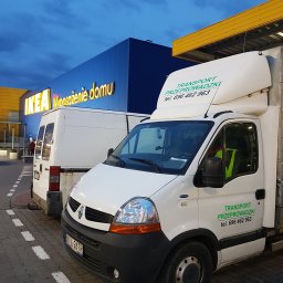 Usługi Transportowe Robert Kołodziejski - Dwa białe busy transportowe zaparkowane przed budynkiem IKEA, jeden z zielonym napisem 'TRANSPORT PRZEPROWADZKI' i numerem telefonu.