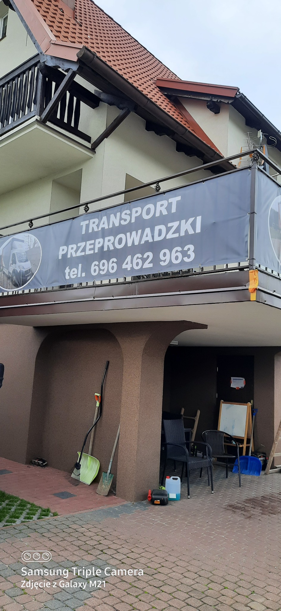 Baner reklamowy firmy transportowej 'Transport Przeprowadzki' z numerem telefonu, umieszczony na balkonie budynku mieszkalnego, z widocznymi narzędziami ogrodniczymi i meblami ogrodowymi...