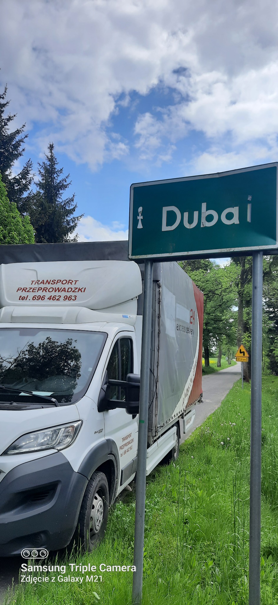Biały bus transportowy z napisem 'TRANSPORT PRZEPROWADZKI' zaparkowany obok drogowskazu wskazującego kierunek na Dubai, na tle zieleni i błękitnego nieba z chmurami.