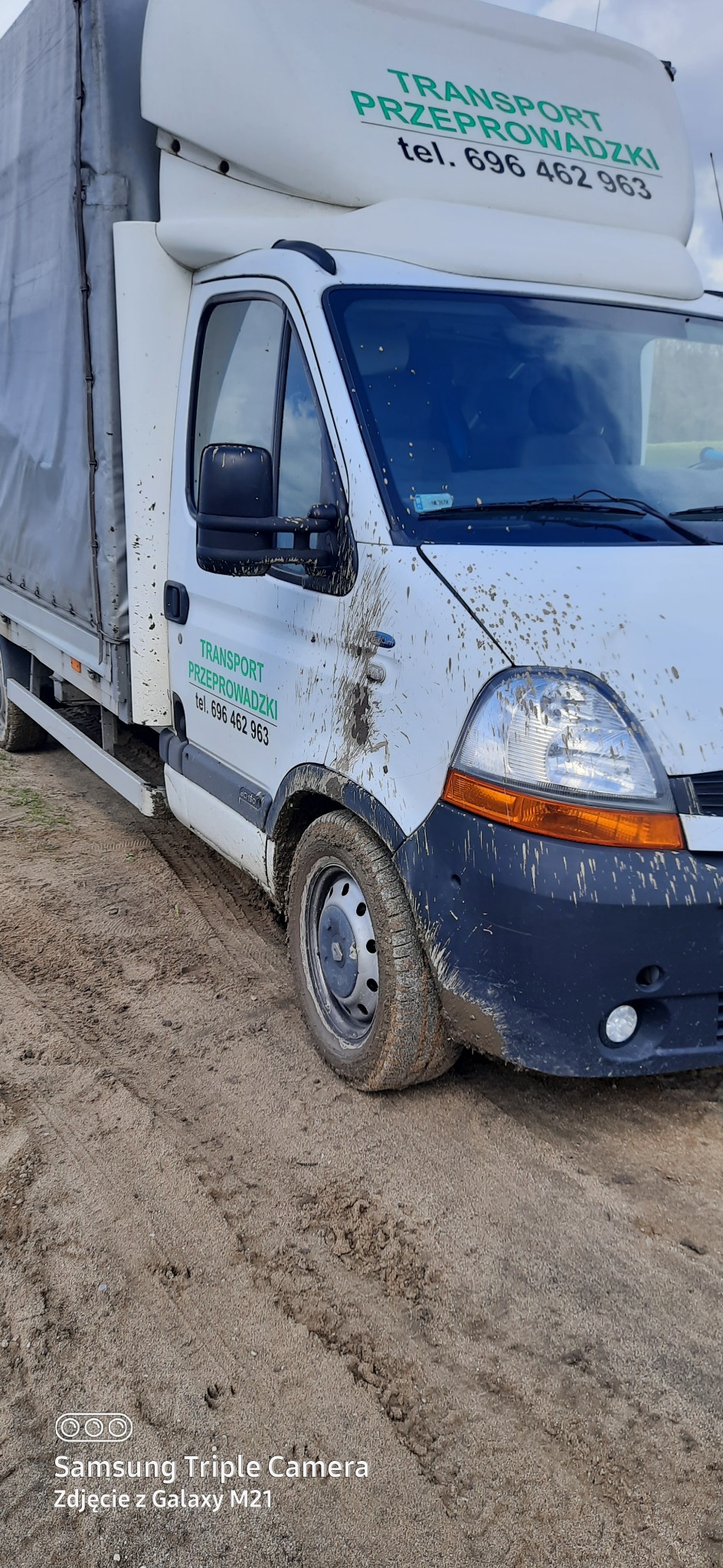 Biały bus transportowy z zielonym napisem 'TRANSPORT PRZEPROWADZKI' mocno zabrudzony błotem, stojący na piaszczystym podłożu.