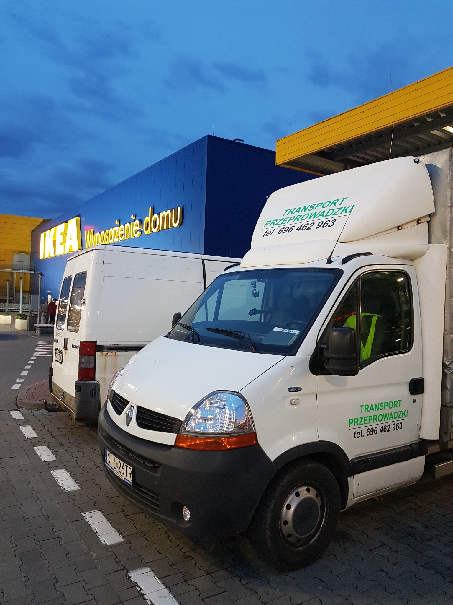 Dwa białe busy transportowe zaparkowane przed budynkiem IKEA, jeden z zielonym napisem 'TRANSPORT PRZEPROWADZKI' i numerem telefonu.