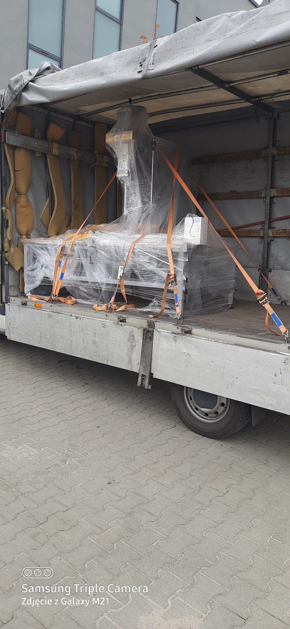 Maszyna przemysłowa owinięta folią stretch, zabezpieczona pomarańczowymi pasami transportowymi w przestrzeni ładunkowej busa z plandeką.