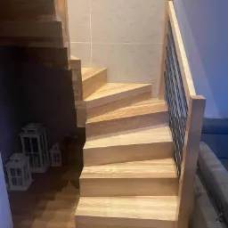 Jasne, drewniane schody z metalową balustradą w nowoczesnym wnętrzu. Widoczne lampiony dekoracyjne pod schodami i fragment szarej sofy w tle.