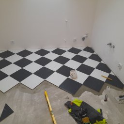 Adam Wiśniewski - Podłoga w szachownicę z białych i czarnych płytek w trakcie układania w pomieszczeniu z białymi ścianami, widoczne narzędzia glazurnicze, poziomica i odzież robocza.