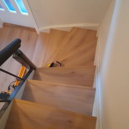 Adam Wiśniewski - Widok z góry na drewniane schody w Mogilnie podczas montażu, widoczne narzędzia, słuchawki ochronne i fragment obuwia montera.