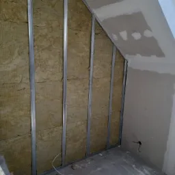 Skośna ściana poddasza z metalowym stelażem wypełnionym wełną mineralną, częściowo pokryta płytami, widoczna drabina aluminiowa z narzędziami.
