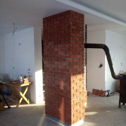 Adam Wiśniewski - Wykończona cegłą klinkierową kolumna w surowym wnętrzu, z widocznym fragmentem komina i przygotowanymi cegłami na podłodze, oświetlona naturalnym światłem padającym z boku.