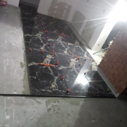 Adam Wiśniewski - Ciemna, marmurowa płytka podłogowa w trakcie montażu, widoczne pomarańczowe krzyżyki dystansowe, częściowo położona, z niedokończonymi krawędziami i świeżym cementem, nieostre tło z fragmentem...