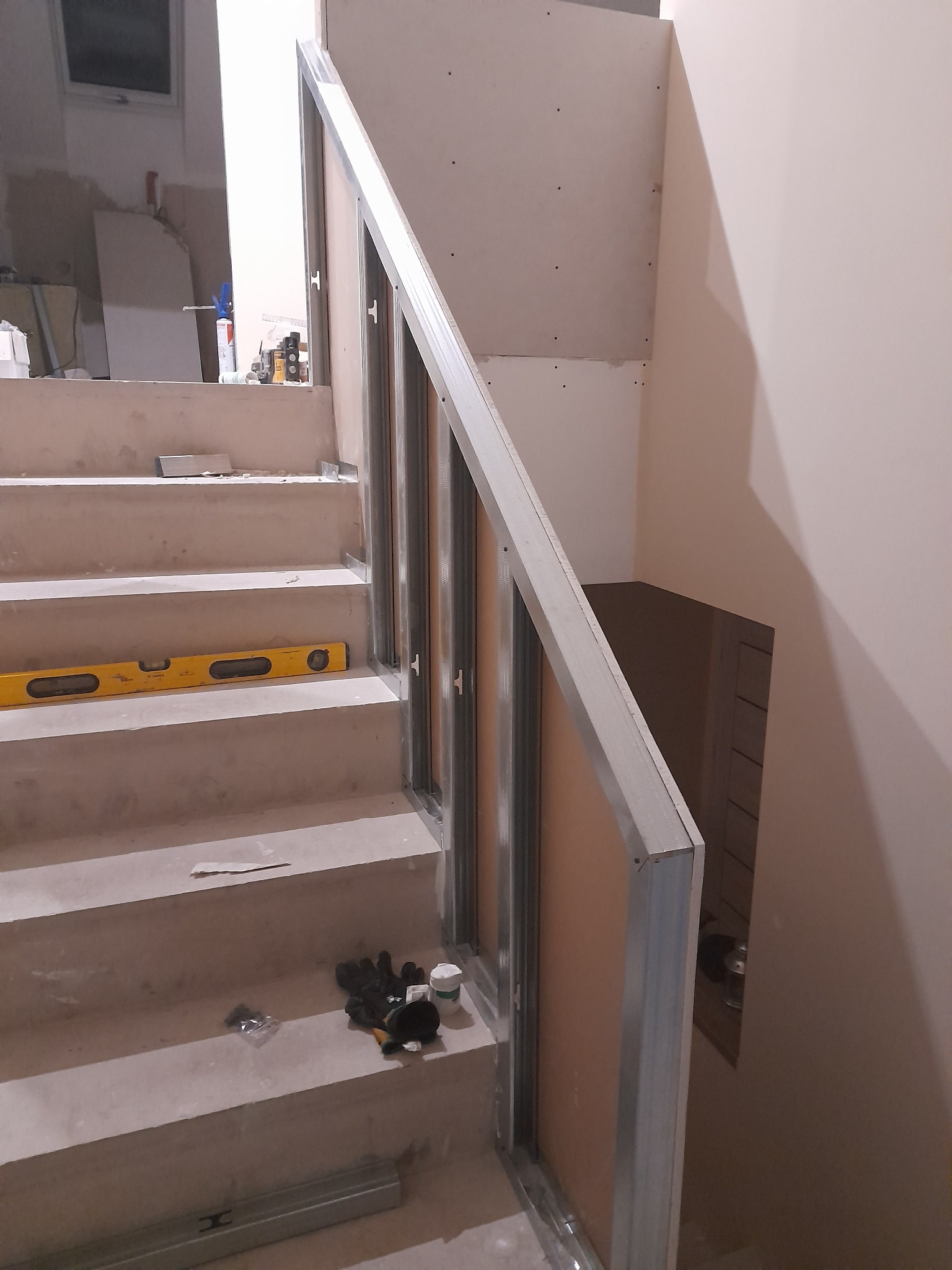 Montaż ściany z profili i płyt na schodach, poziomica leży na stopniu, widoczne rękawice robocze i puszka z masą szpachlową.