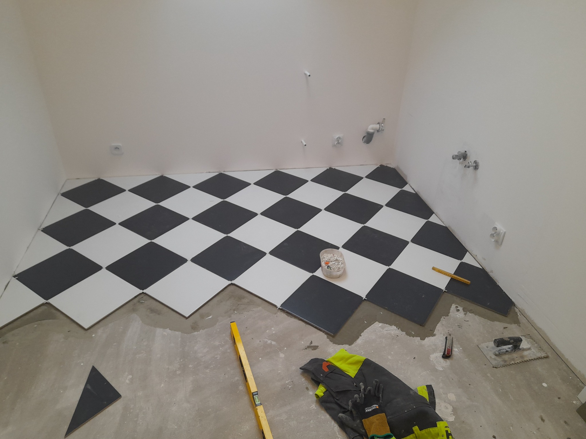Podłoga w szachownicę z białych i czarnych płytek w trakcie układania w pomieszczeniu z białymi ścianami, widoczne narzędzia glazurnicze, poziomica i odzież robocza.