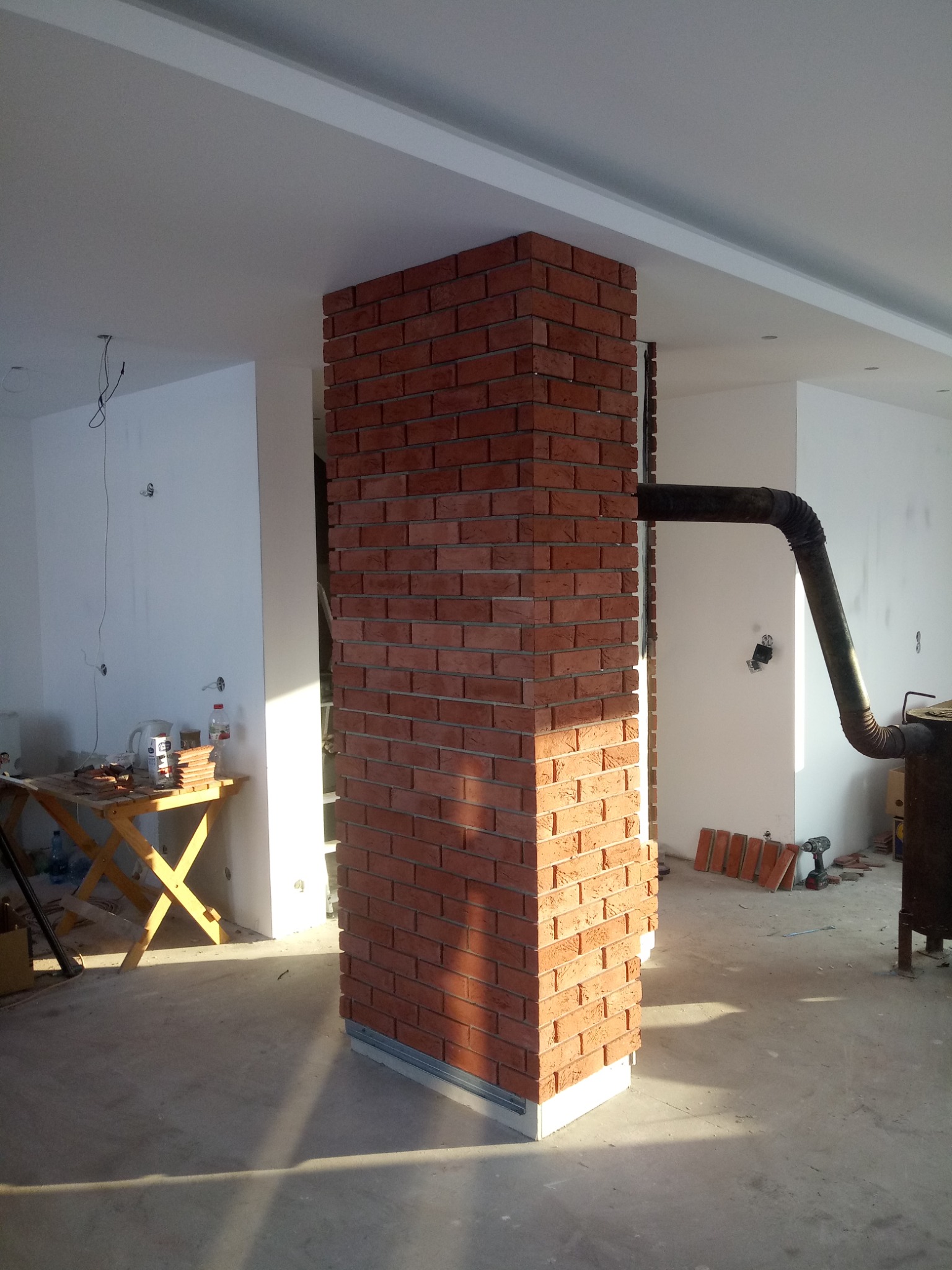 Wykończona cegłą klinkierową kolumna w surowym wnętrzu, z widocznym fragmentem komina i przygotowanymi cegłami na podłodze, oświetlona naturalnym światłem padającym z boku.