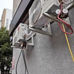 JDGKlima - Dwa agregaty klimatyzacji LG Dual Inverter zamontowane na szarej ścianie budynku, podłączone do manometrów i przewodów serwisowych, widoczne krople wody na obudowie, sugerujące wilgotną pogodę.