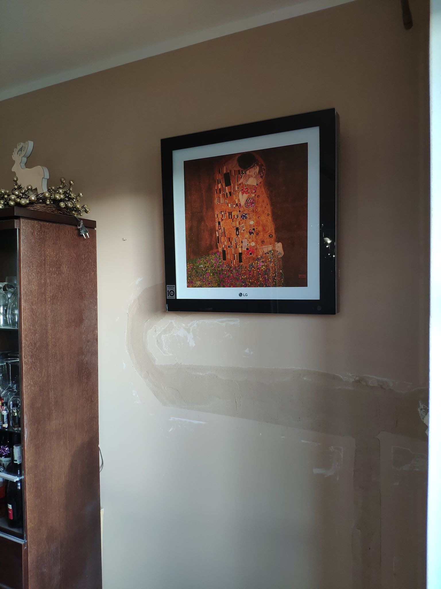 Telewizor LG zawieszony na ścianie z widocznymi śladami po instalacji, prezentujący obraz Gustava Klimta 'Pocałunek', obok drewniana witryna z dekoracją świąteczną.