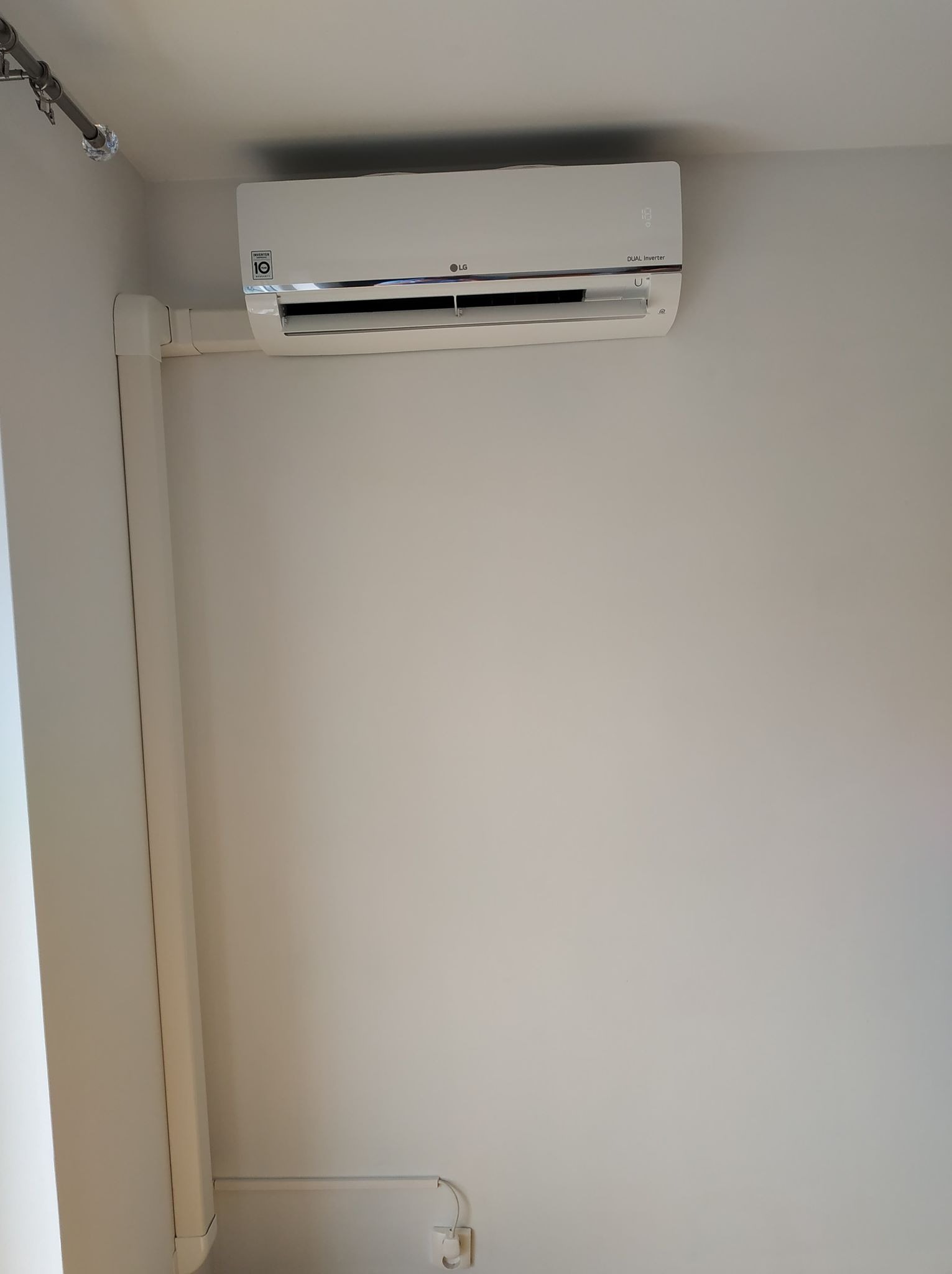 Biały klimatyzator LG Dual Inverter zamontowany na jasnej ścianie, z widocznymi elementami instalacji elektrycznej i rurami odprowadzającymi skropliny.