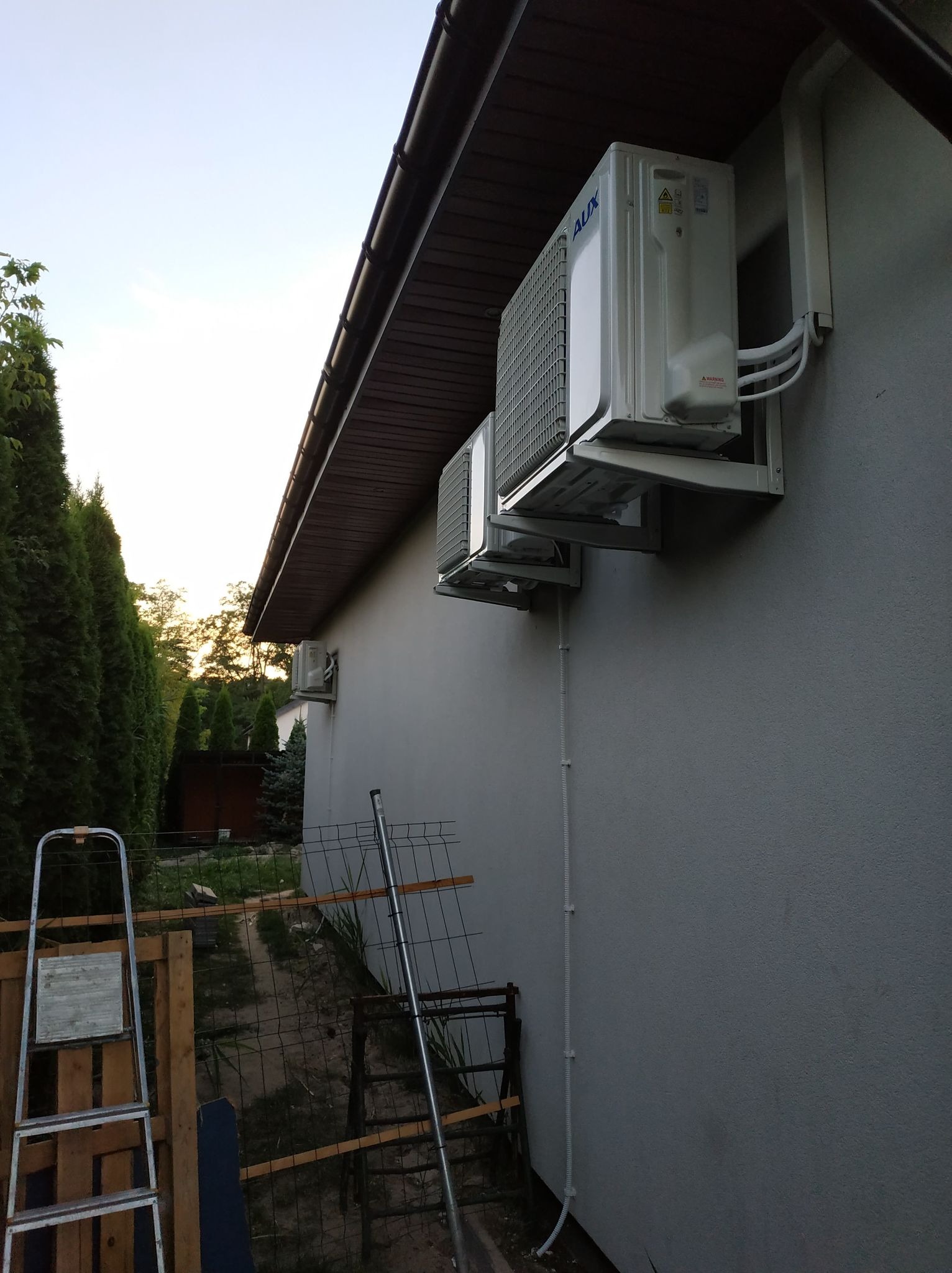 Dwa klimatyzatory marki AUX zamontowane na ścianie budynku w Warszawie, widoczne rury odprowadzające, drabina oparta o ogrodzenie w tle.