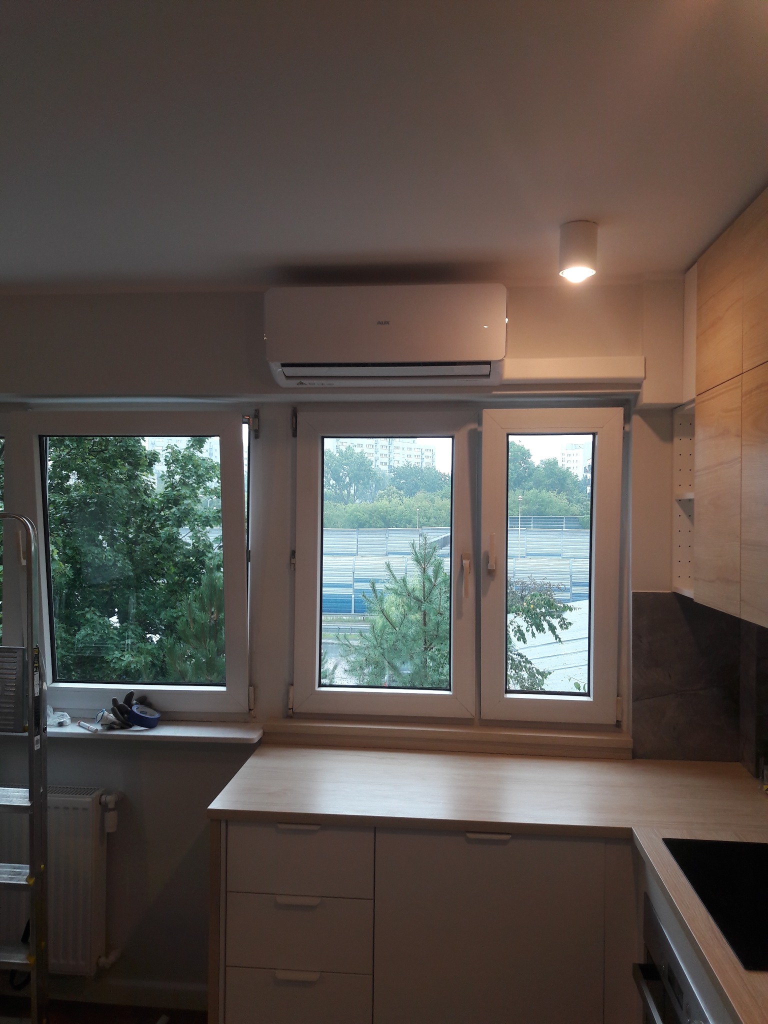 Biały klimatyzator zamontowany nad oknem w jasnym wnętrzu kuchni, widok na zieleń za oknem, drabina oparta o ścianę po lewej stronie.