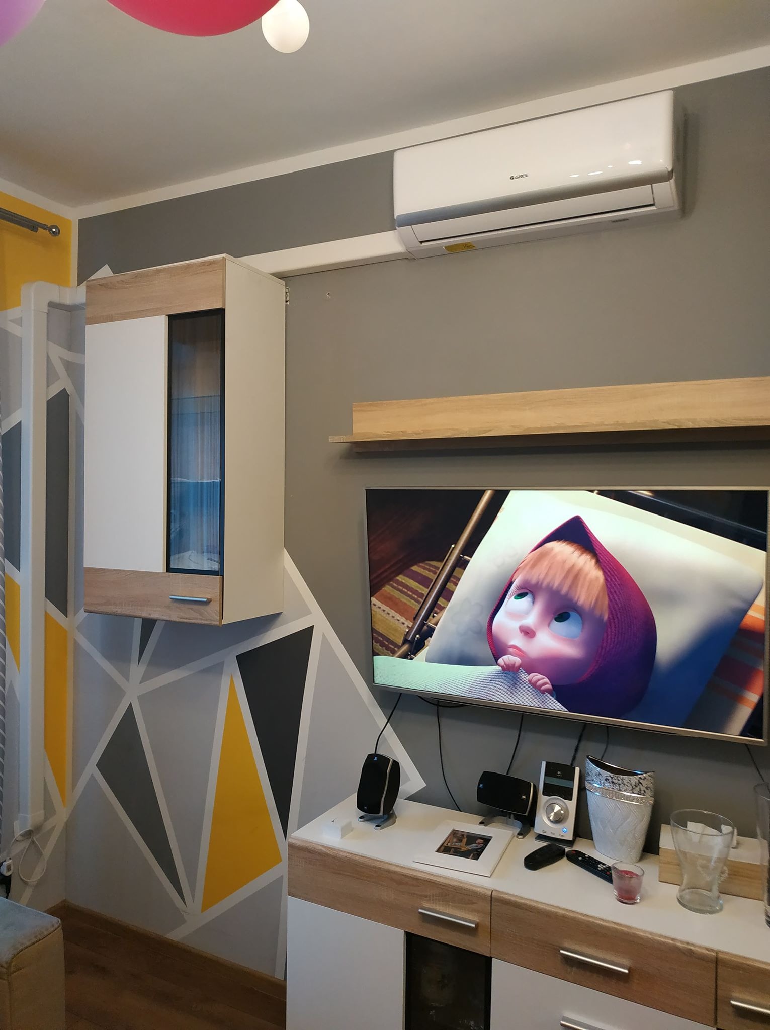 Biała jednostka wewnętrzna zamontowana na szarej ścianie w pokoju z telewizorem, szafką RTV i geometrycznym wzorem na ścianie. Widoczny model Gree.
