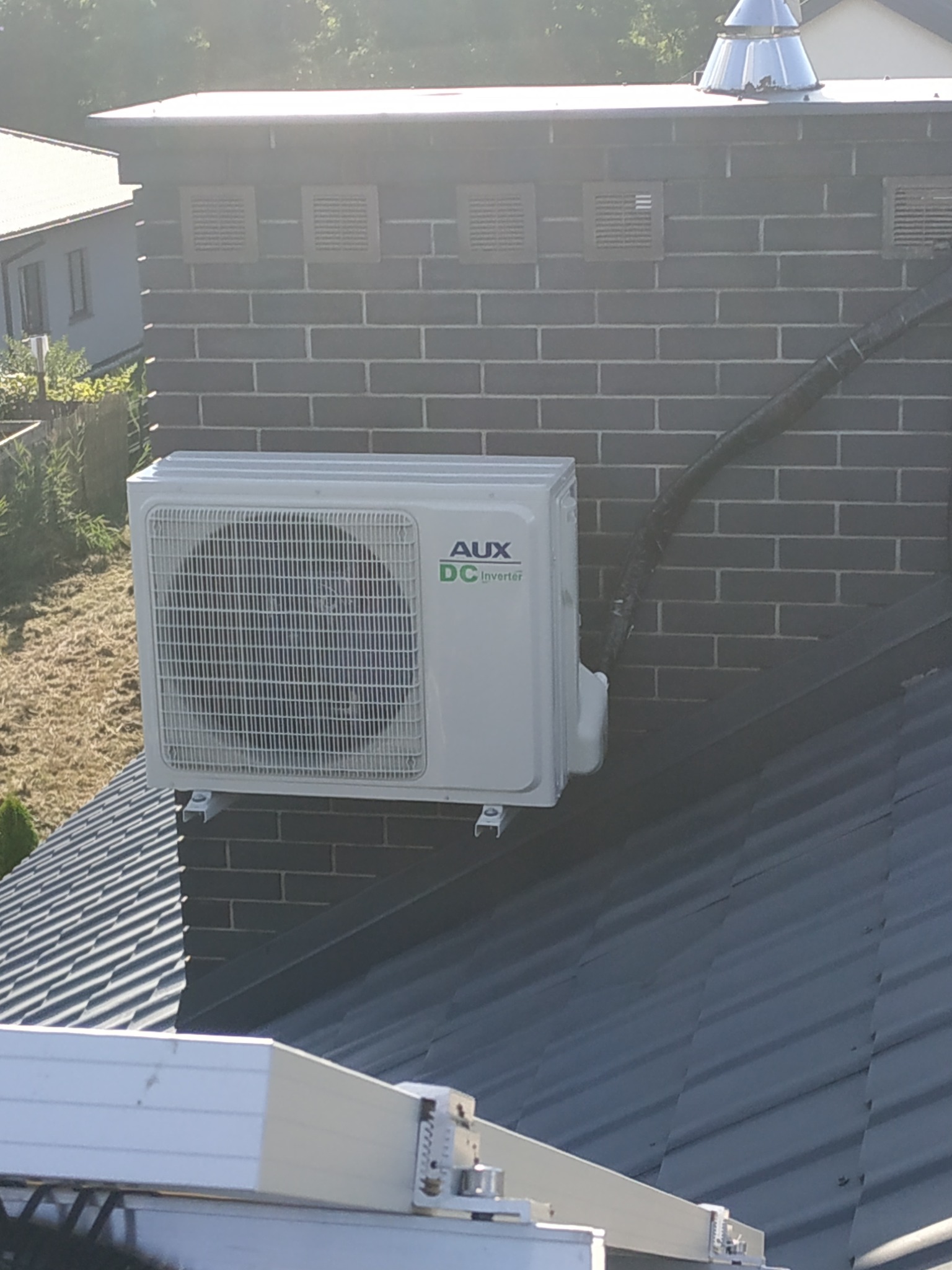 Zewnętrzna jednostka klimatyzatora AUX DC Inverter zamontowana na ścianie komina z szarej cegły, widok z dachu pokrytego blachą trapezową.