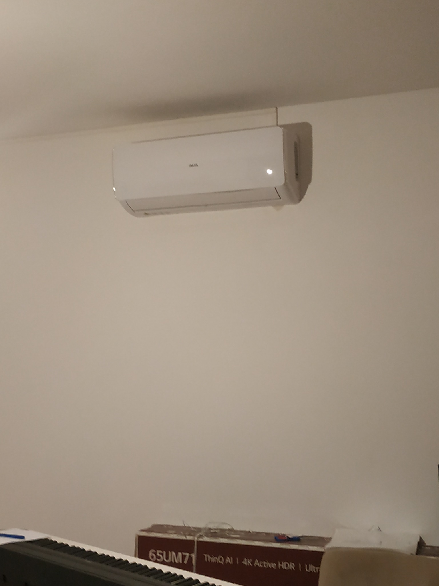Biały klimatyzator marki AUX zamontowany na ścianie w pokoju, widoczna dioda sygnalizacyjna, pod nim karton po telewizorze i fragment pianina.