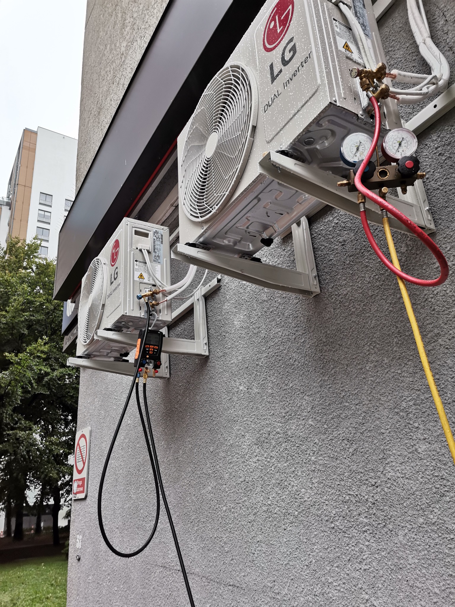 Dwa agregaty klimatyzacji LG Dual Inverter zamontowane na szarej ścianie budynku, podłączone do manometrów i przewodów serwisowych, widoczne krople wody na obudowie, sugerujące wilgotną pogodę.