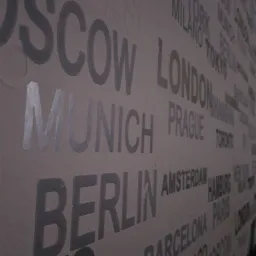 Ściana pokryta tapetą z motywem nazw miast w odcieniach szarości, widoczne napisy: Moskwa, Monachium, Berlin, Londyn, Praga, Amsterdam, Barcelona, Chicago.