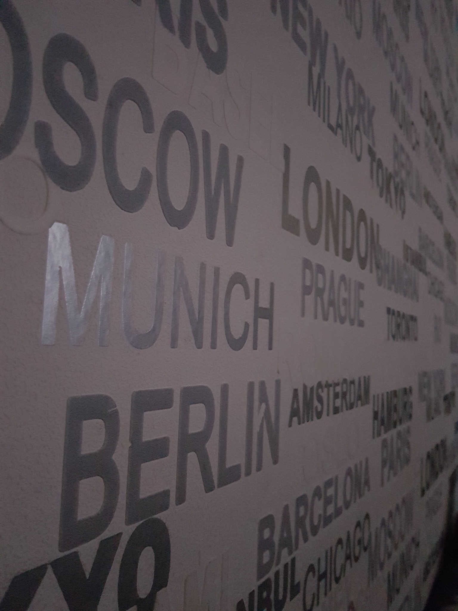 Ściana pokryta tapetą z motywem nazw miast w odcieniach szarości, widoczne napisy: Moskwa, Monachium, Berlin, Londyn, Praga, Amsterdam, Barcelona, Chicago.