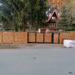 Nowoczesne ogrodzenie z jasnego drewna z czarną metalową bramą wjazdową, w tle dom z drewnianą elewacją i zielone drzewa, przy drodze biała torba z resztkami materiał&oacute;w budowlanych.