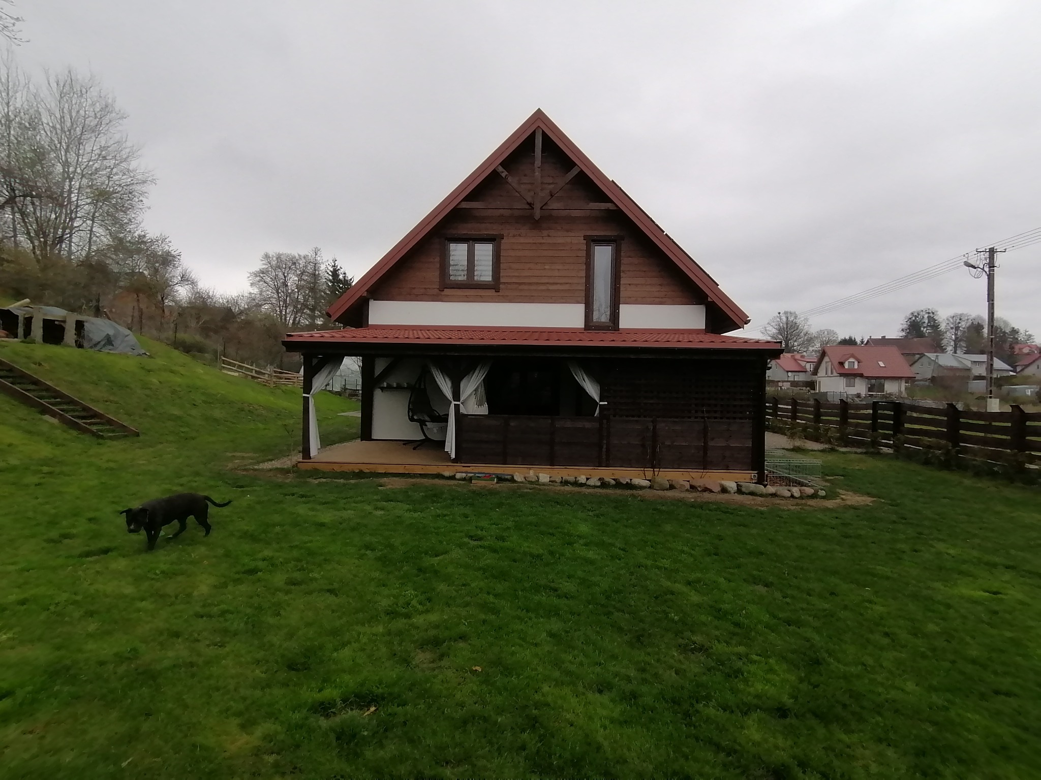Drewniany dom z tarasem z ciemnego drewna i białymi zasłonami, na trawniku czarny pies, w tle widoczne inne domy.