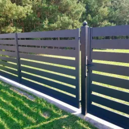 Nowoczesne ogrodzenie panelowe z poziomych, szarych profili, zakończone kulistymi elementami na słupkach, otwierana furtka, widok na trawnik i drzewa.