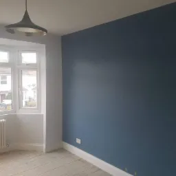 Pomieszczenie po malowaniu: ściana w odcieniu głębokiego granatu, białe okno wykuszowe z widokiem na domy, grzejnik pod oknem, drewniana podłoga przed cyklinowaniem, nowoczesne lampy sufitowe.