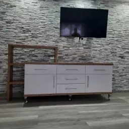 Biała komoda z drewnianym blatem i regałem, ustawiona na tle ściany z szarej cegły, z telewizorem wiszącym powyżej.